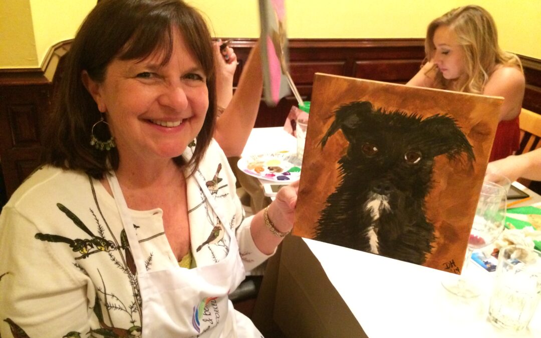 Brown haired woman holds up painting of her black and white dog.